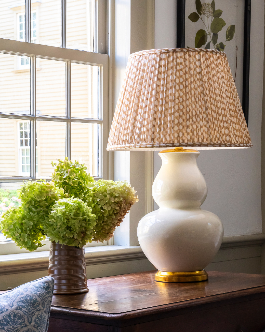 White lamp with a textured beige shade on a wooden table next to a vase of green hydrangeas, with a window in the background.