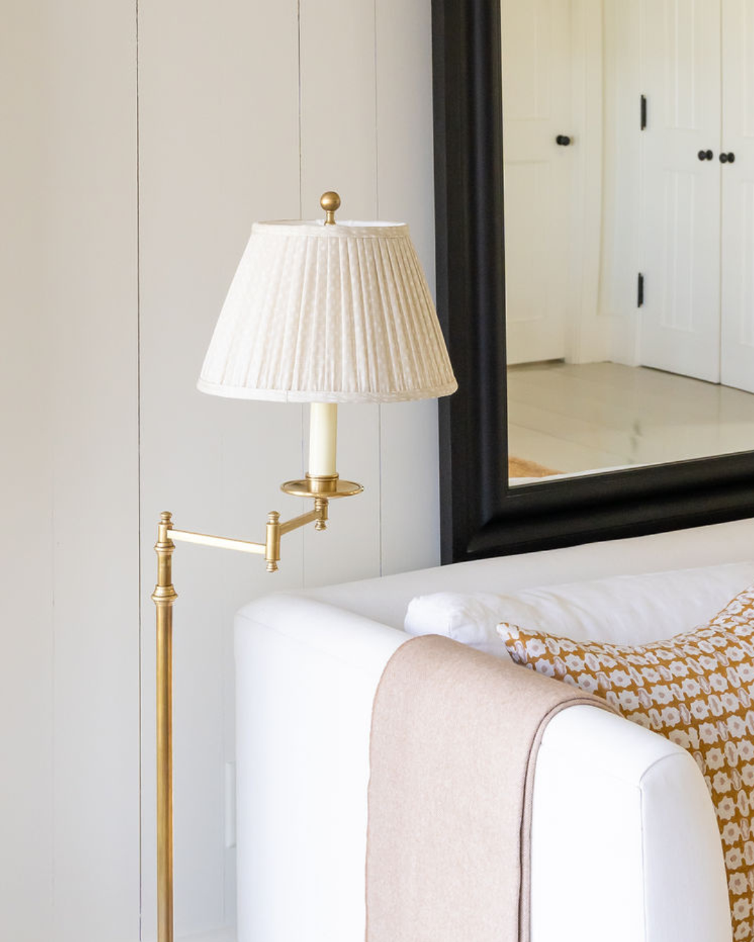 Gold swing-arm wall lamp with pleated white shade next to a white sofa and large mirror.