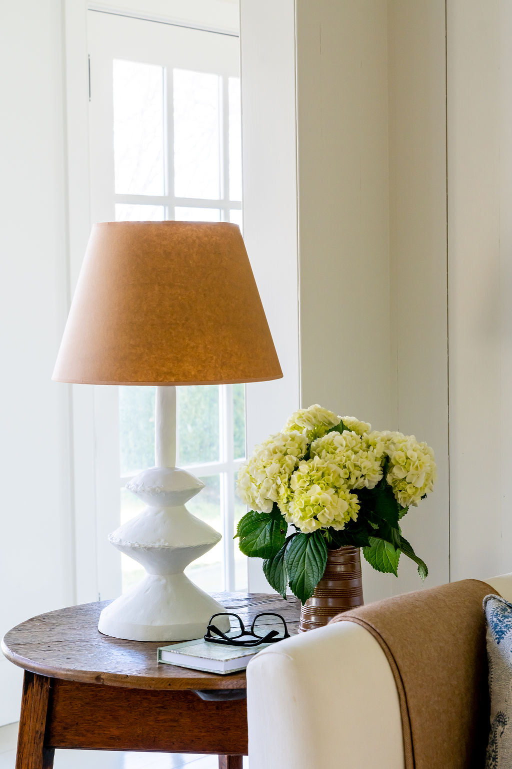 Lamp with brown shade on a table next to a vase of flowers in a room with a window.