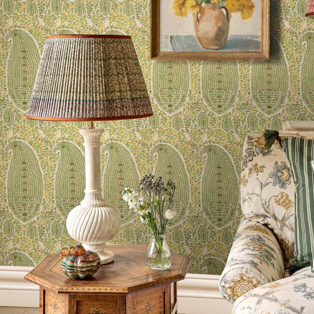A styled interior corner showing a table lamp with a pleated patterned Penny Morrison lampshade and white ribbed base on a wooden side table, set against green floral wallpaper. Part of a floral armchair and framed wall art are visible.