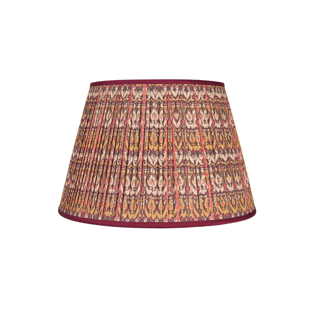 A pleated fabric lampshade by Penny Morrison featuring an intricate ikat pattern in shades of orange, brown, beige, and red, finished with a solid deep burgundy trim.