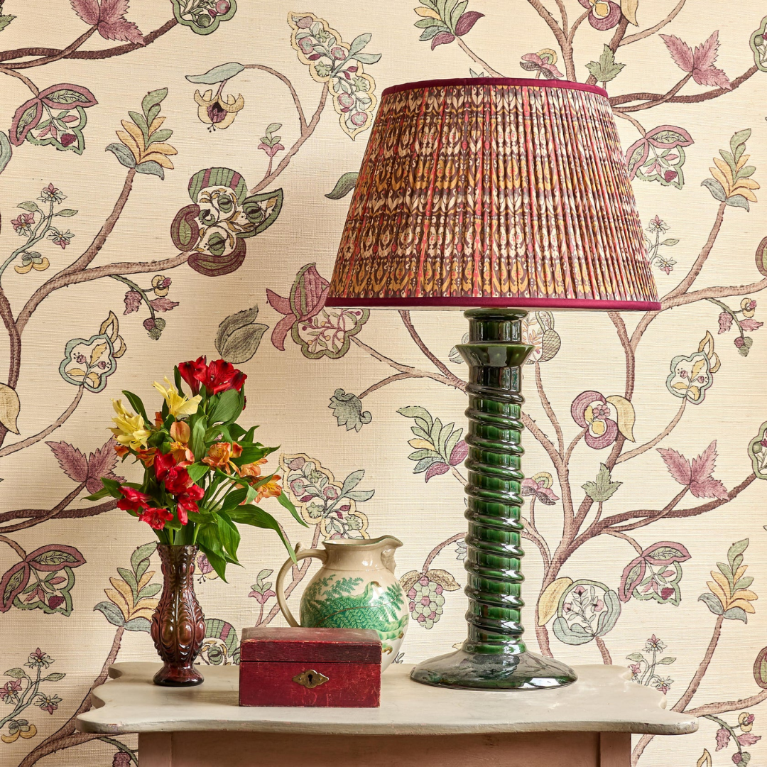 A decorative table lamp with a pleated patterned shade by Penny Morrison in pink, gold, and green, and a ribbed green glass base, styled on a table against a floral wallpaper background.