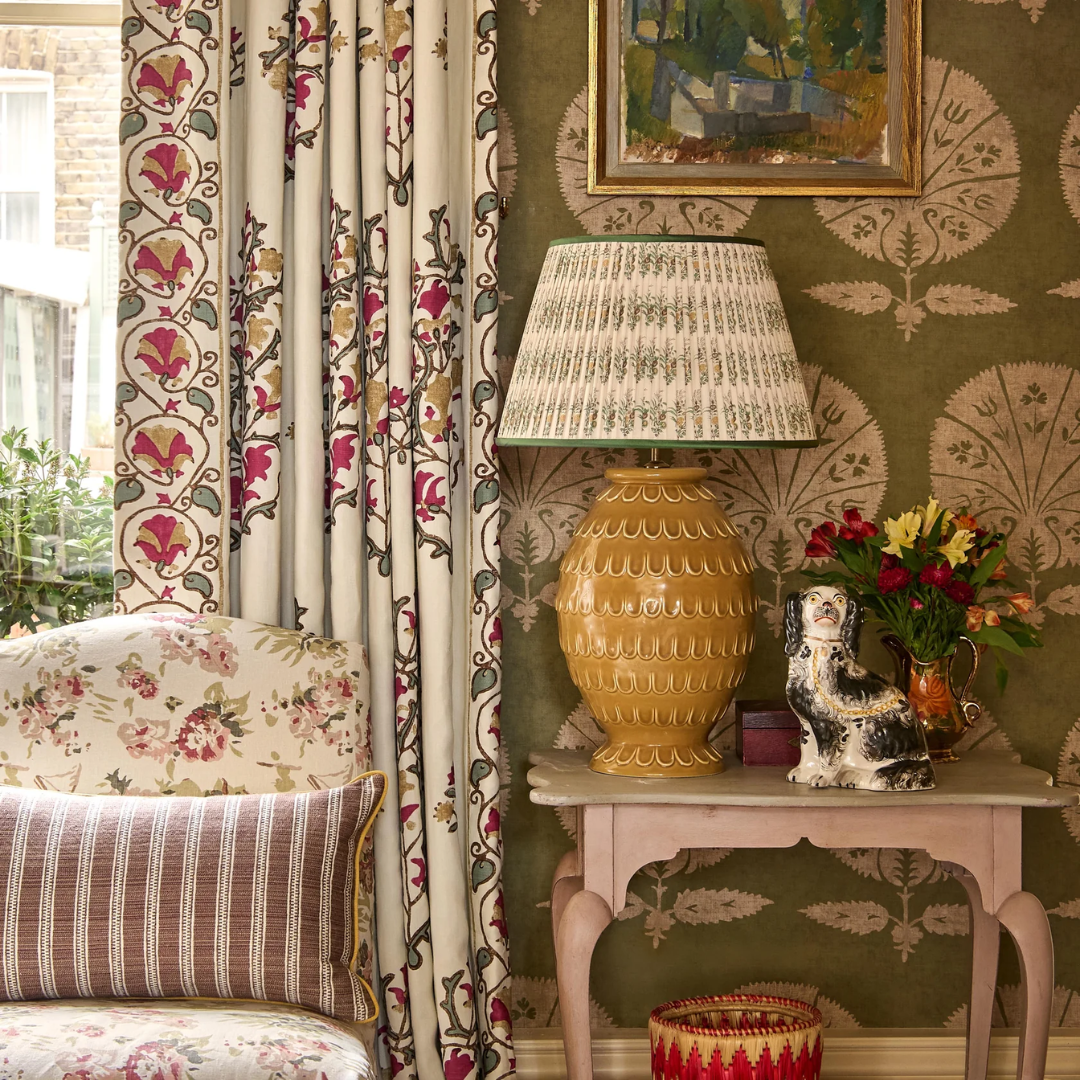 A styled interior corner featuring a pleated lampshade by Penny Morrison, patterned curtains, floral wallpaper, a yellow textured table lamp, a small side table with flowers and a ceramic bird figurine, and part of a matching floral armchair.