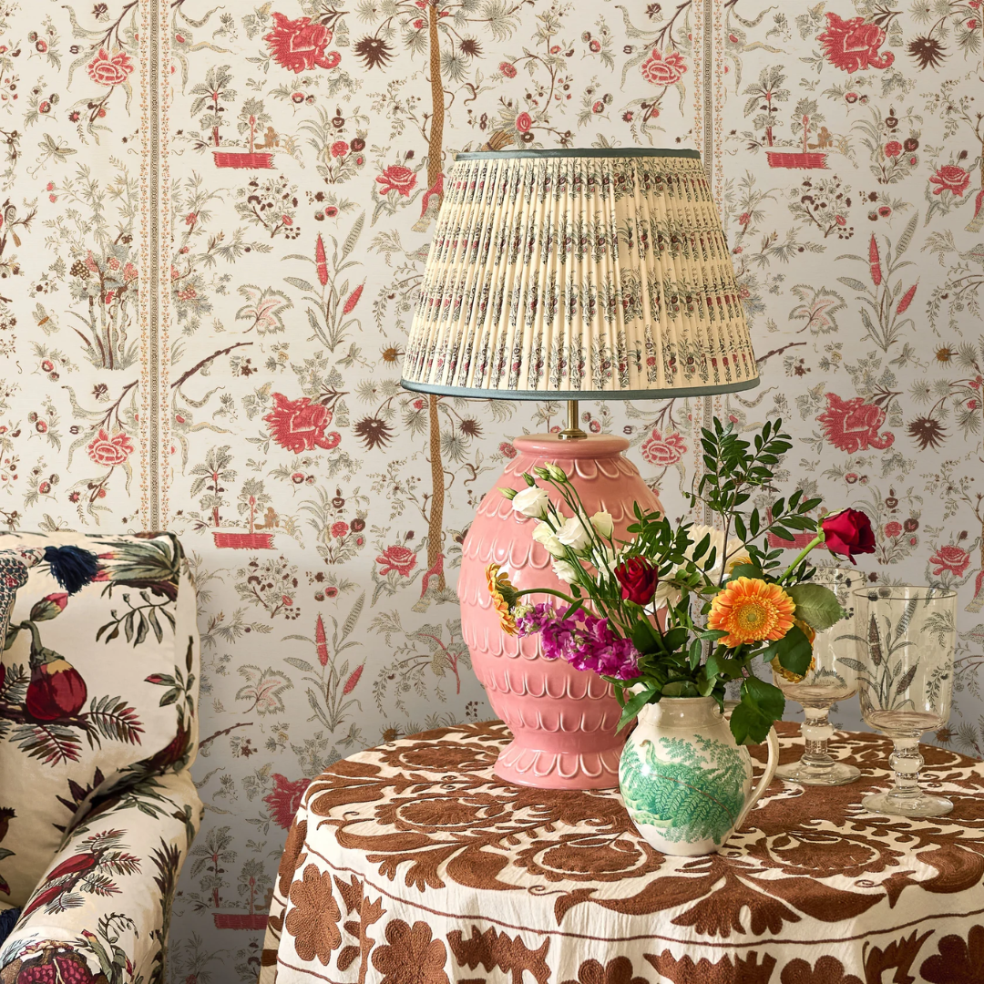 A pink ceramic table lamp with a pleated penny morrison beige and brown patterned shade stands on a round table covered in a brown floral tablecloth. A small green-and-white patterned vase with orange, pink, and white flowers sits beside it against a matching floral wallpaper background.